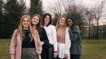 Fünf Frauen stehen nebeneinander im Freien, lächelnd und in unterschiedlichen Outfits. Die Umgebung ist grün mit Bäumen im Hintergrund.