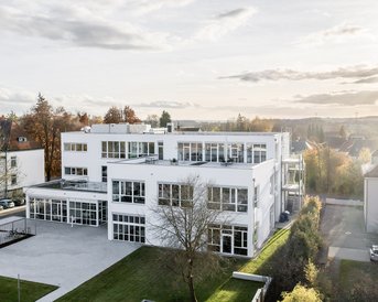 Moderne, mehrstöckige weiße Gebäude mit großen Fenstern und Terrassen, umgeben von Bäumen und Wiese. Der Himmel ist teilweise bewölkt, und es ist Tageslicht.